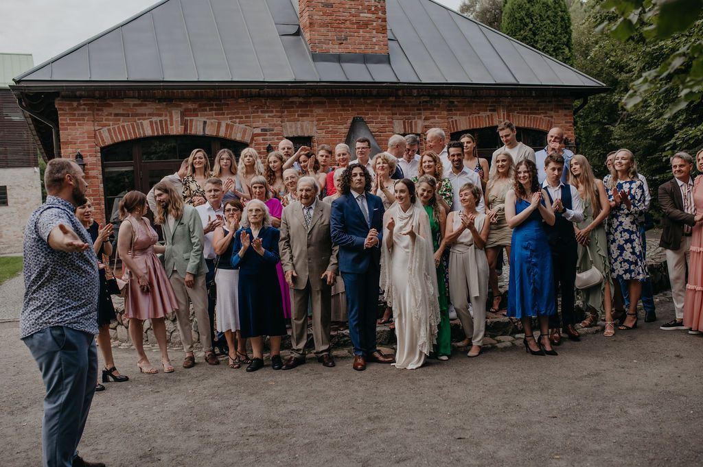 Large wedding group photo with guests applauding outdoors near a rustic brick building.