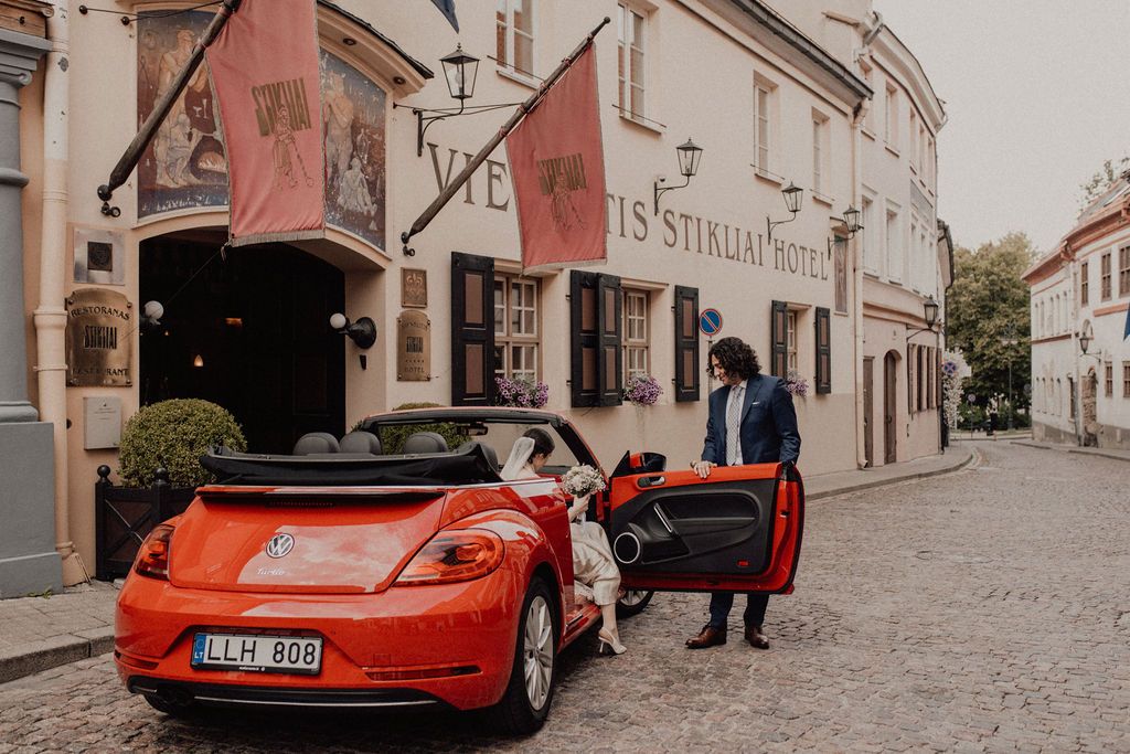 A man opens the door of a red VW Beetle convertible parked on cobblestones outside a historic hotel.