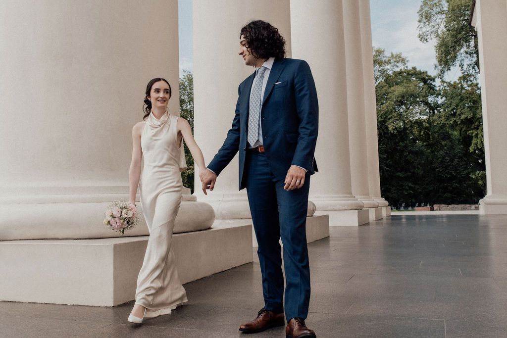Bride in white pantsuit and groom in navy suit hold hands walking between columns at outdoor wedding venue.