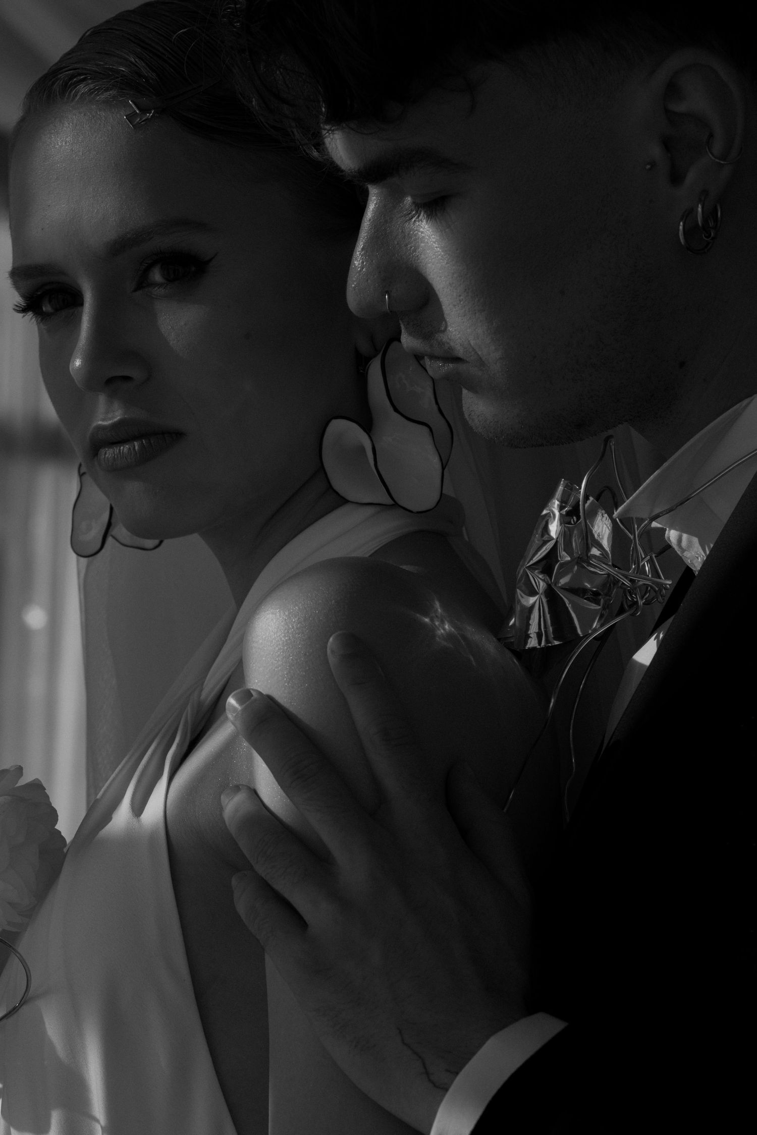 Black and white portrait of an elegant couple, man in suit embracing woman wearing statement earrings and white dress.