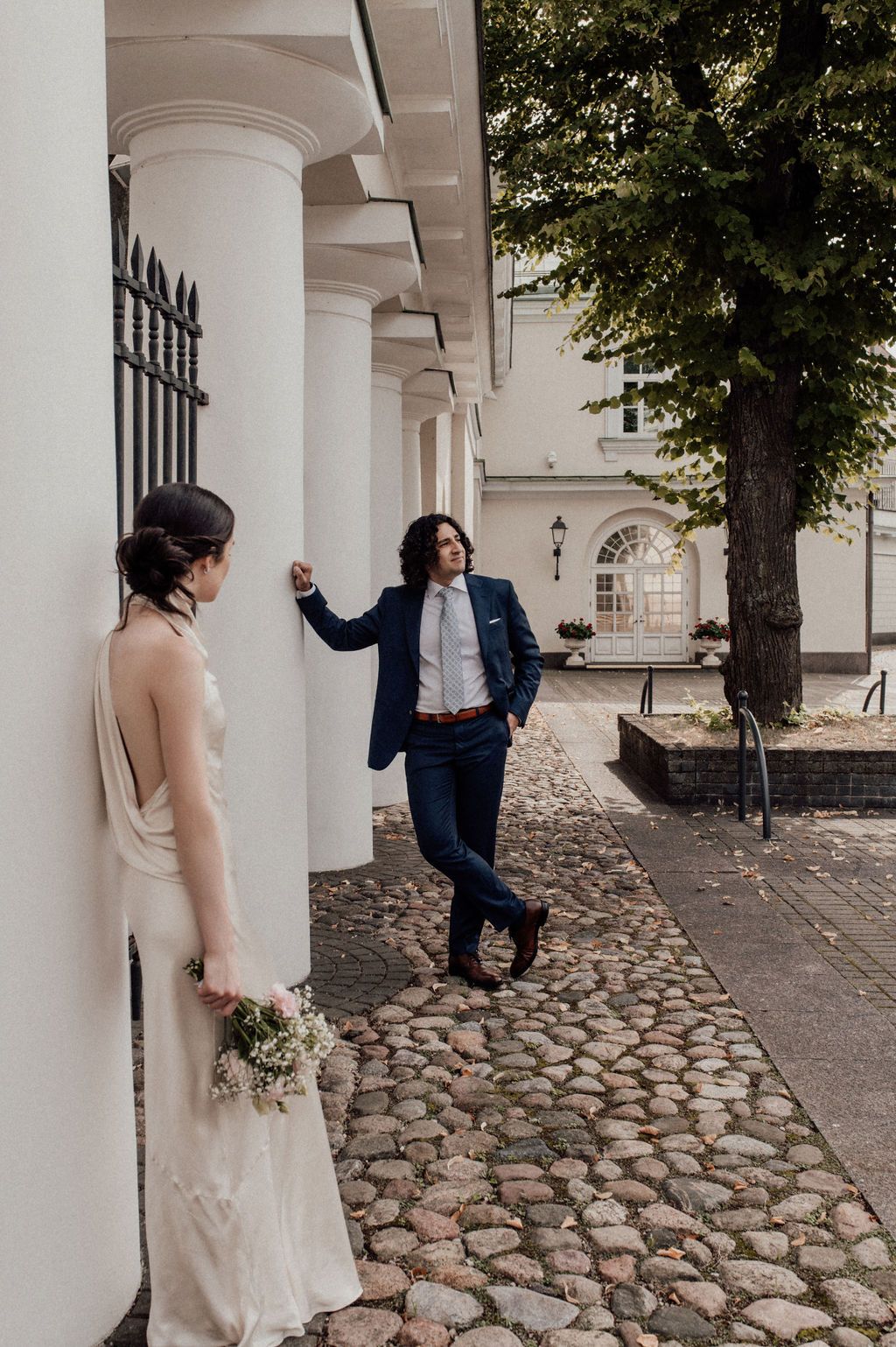 Bride in white dress holds bouquet while groom in navy suit leans against white column on cobblestone path.