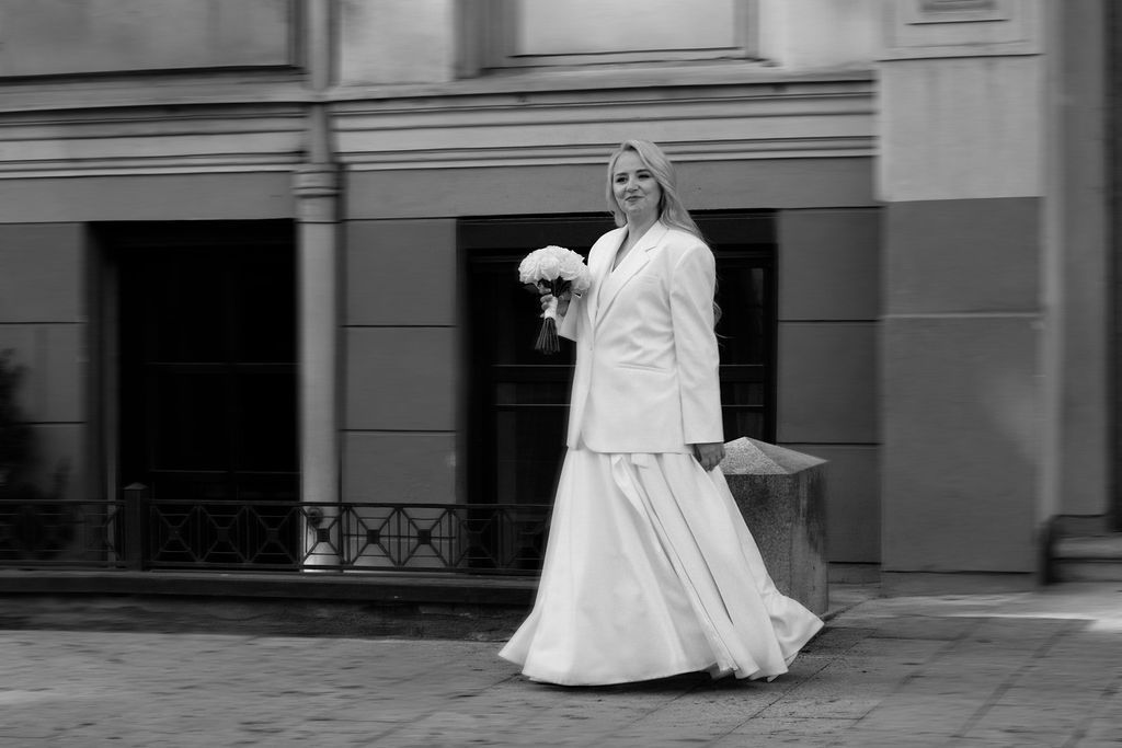 Elegant bride in white gown and jacket holding bouquet, posing outside a classic building in black and white photo.