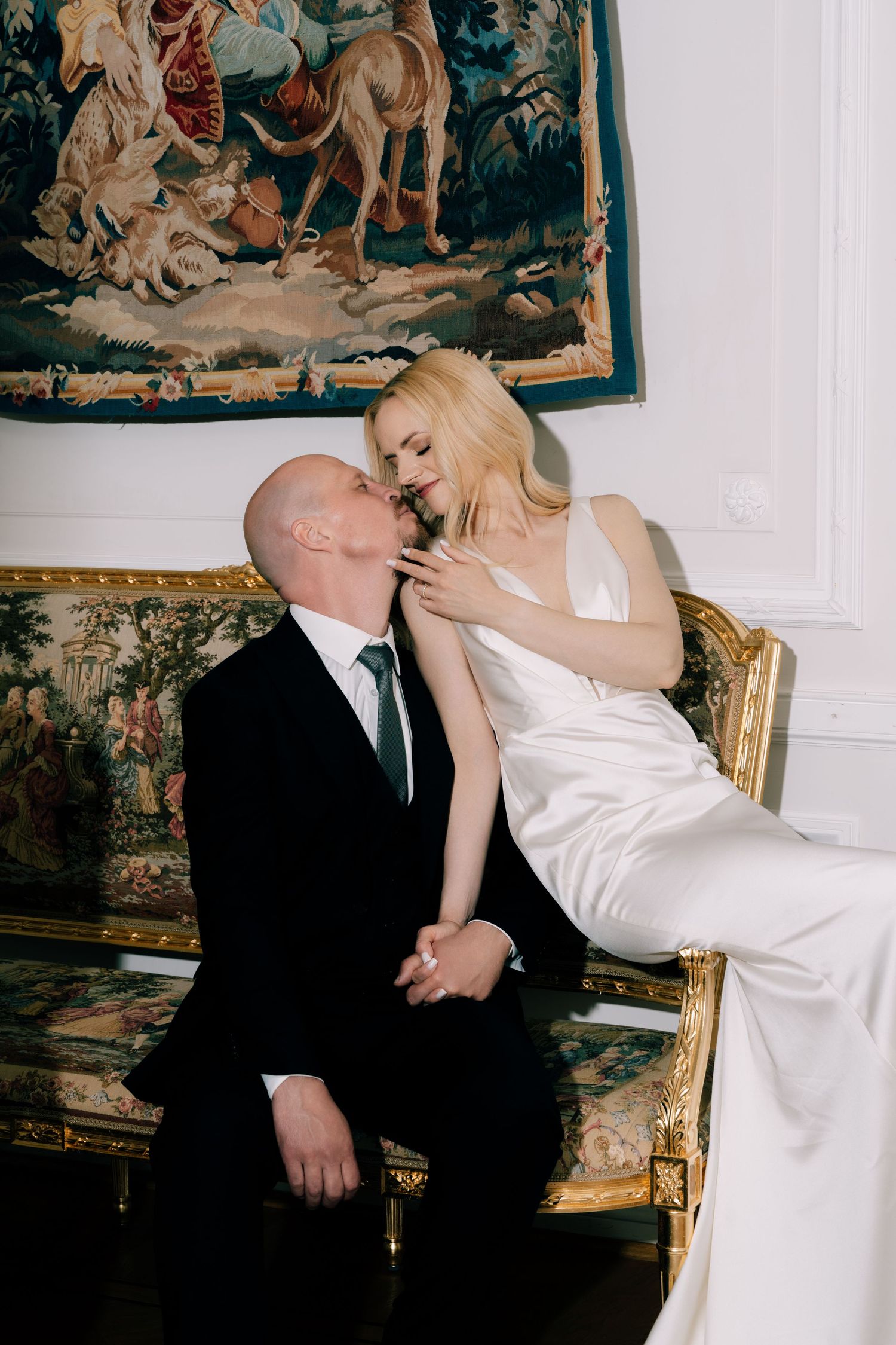 Bride in a white satin wedding dress kisses a groom in a black suit on a gilded antique sofa beneath a tapestry in an elegant room.