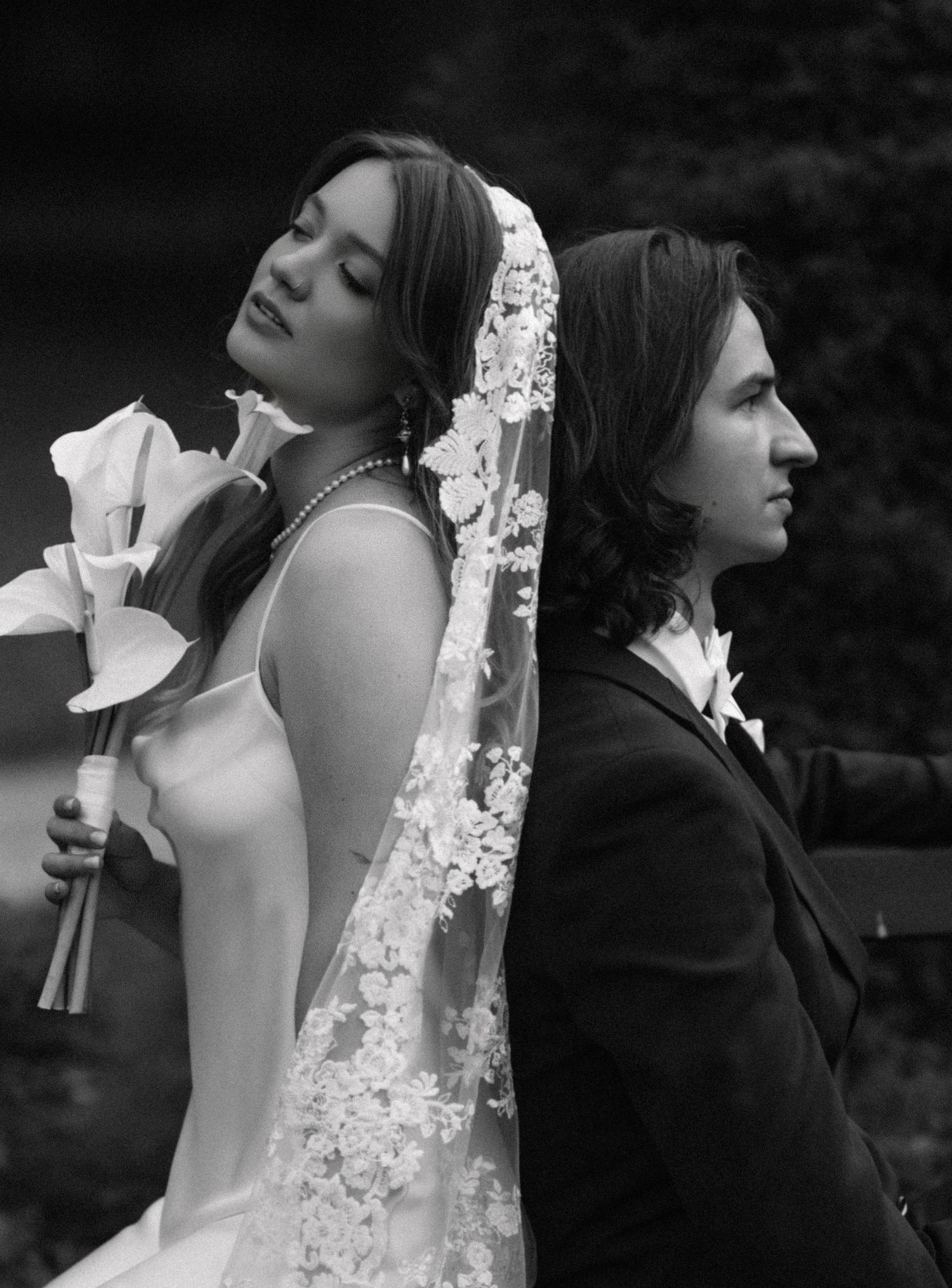 Black and white portrait of a bride in a lace veil and pearl necklace standing back to back with her groom, holding calla lilies.