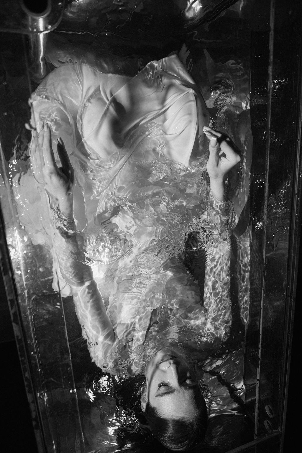 Black and white overhead photo of a woman in a sheer lace gown lying with eyes closed in rippling water inside a glass tank.