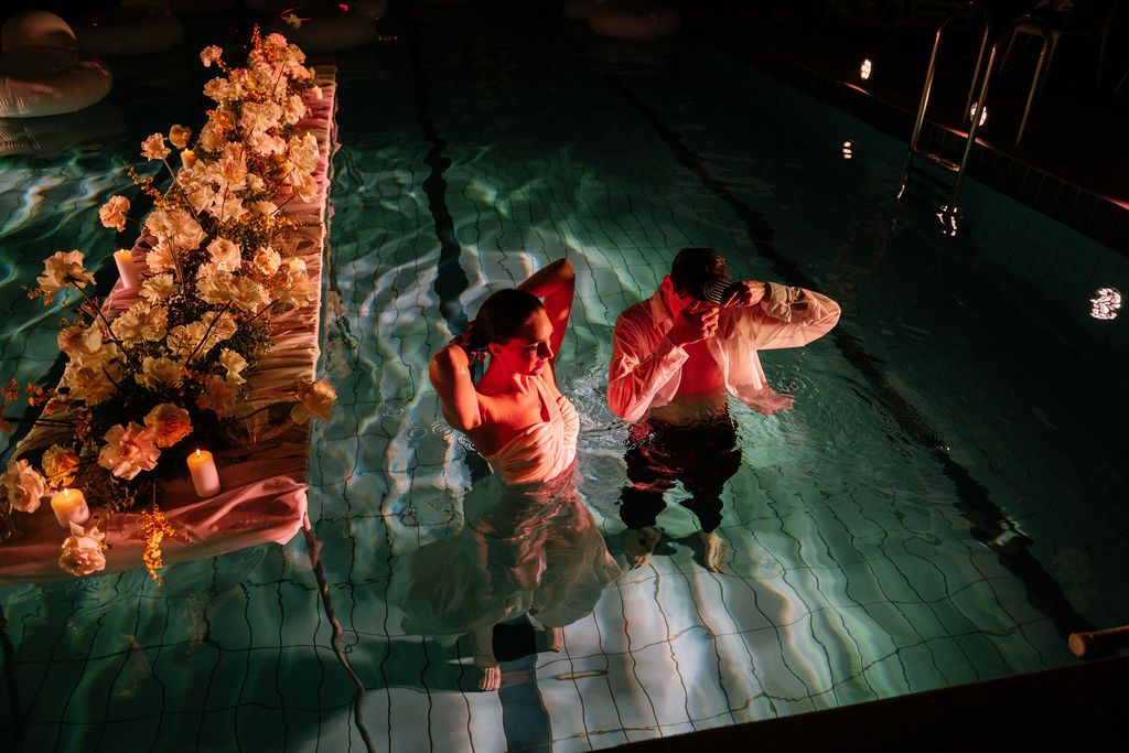 Two people in red tops stand waist-deep in a softly lit pool at night beside a floating arrangement of peach and orange flowers.