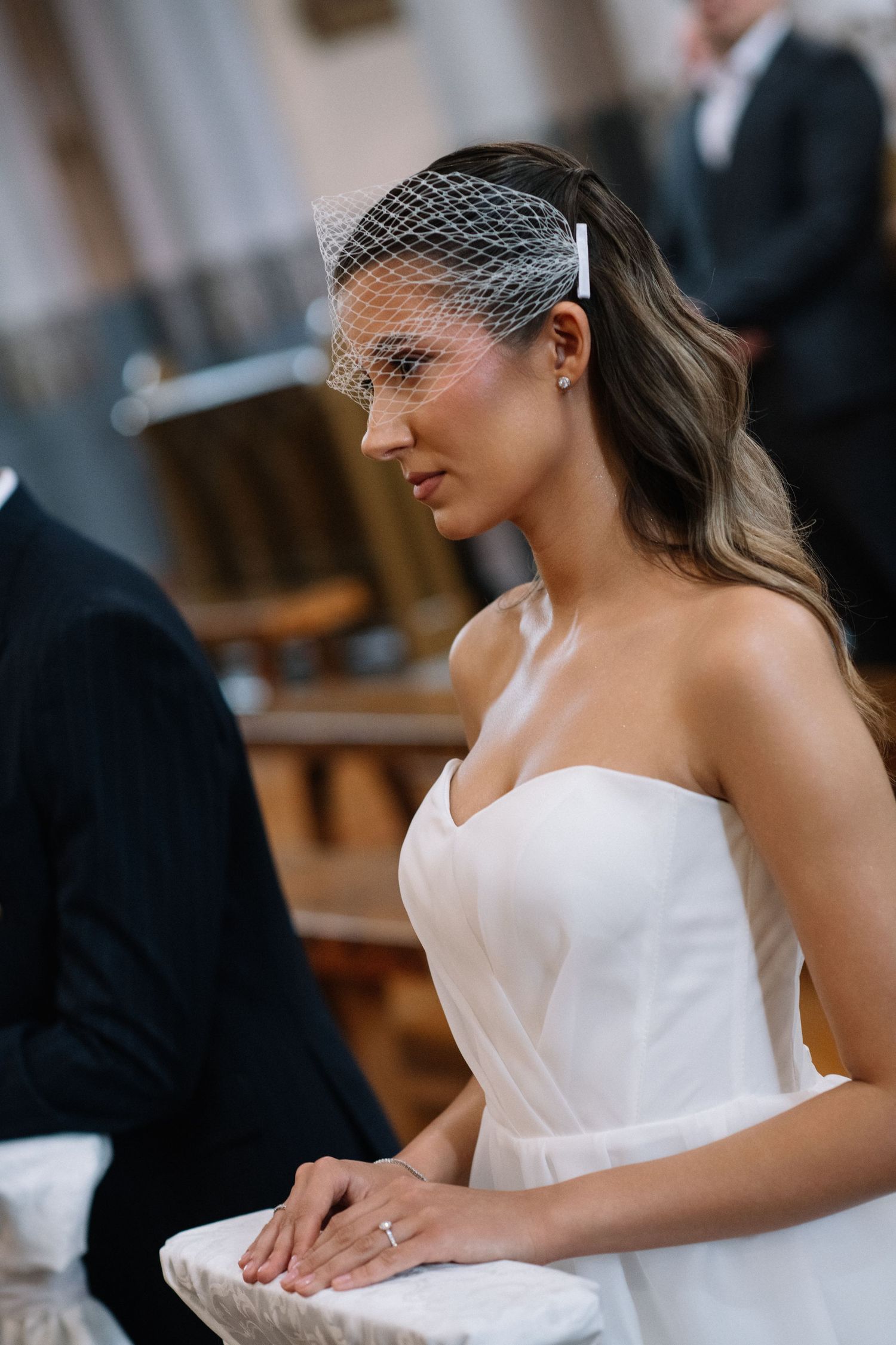 Bride in a white strapless gown and birdcage veil with hair clip kneels with hands folded during a church wedding ceremony.