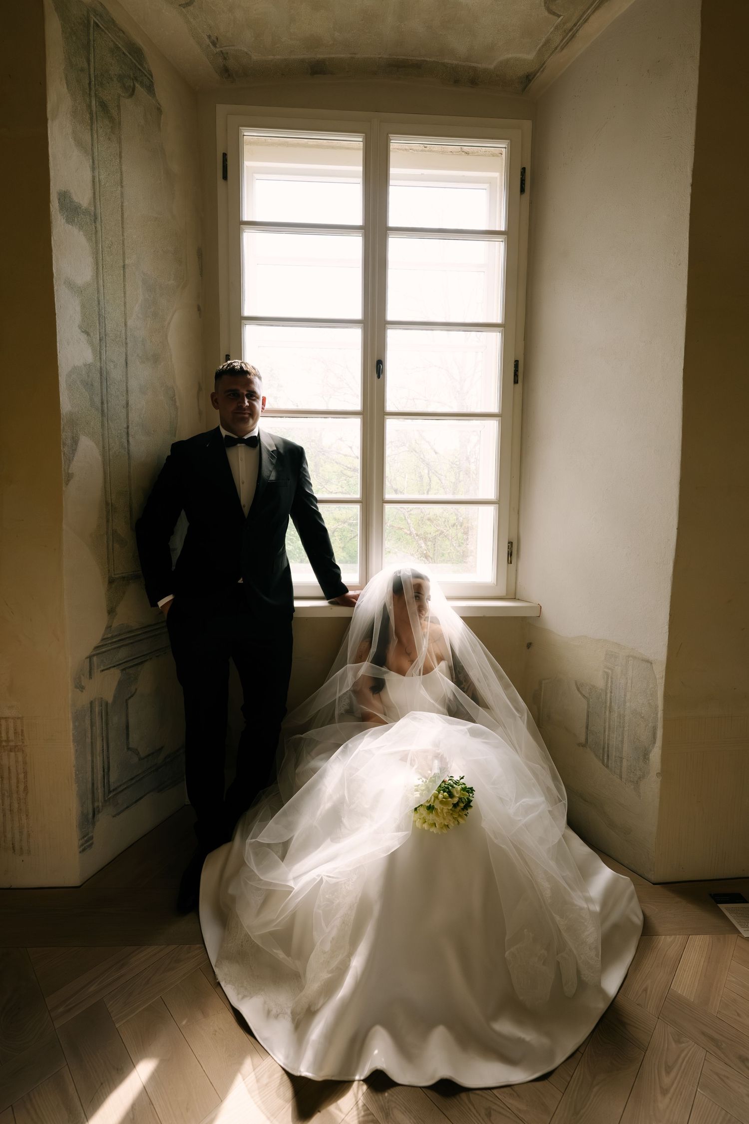A groom in a black suit and bow tie stands by a tall window beside his seated bride in a voluminous white gown and veil.