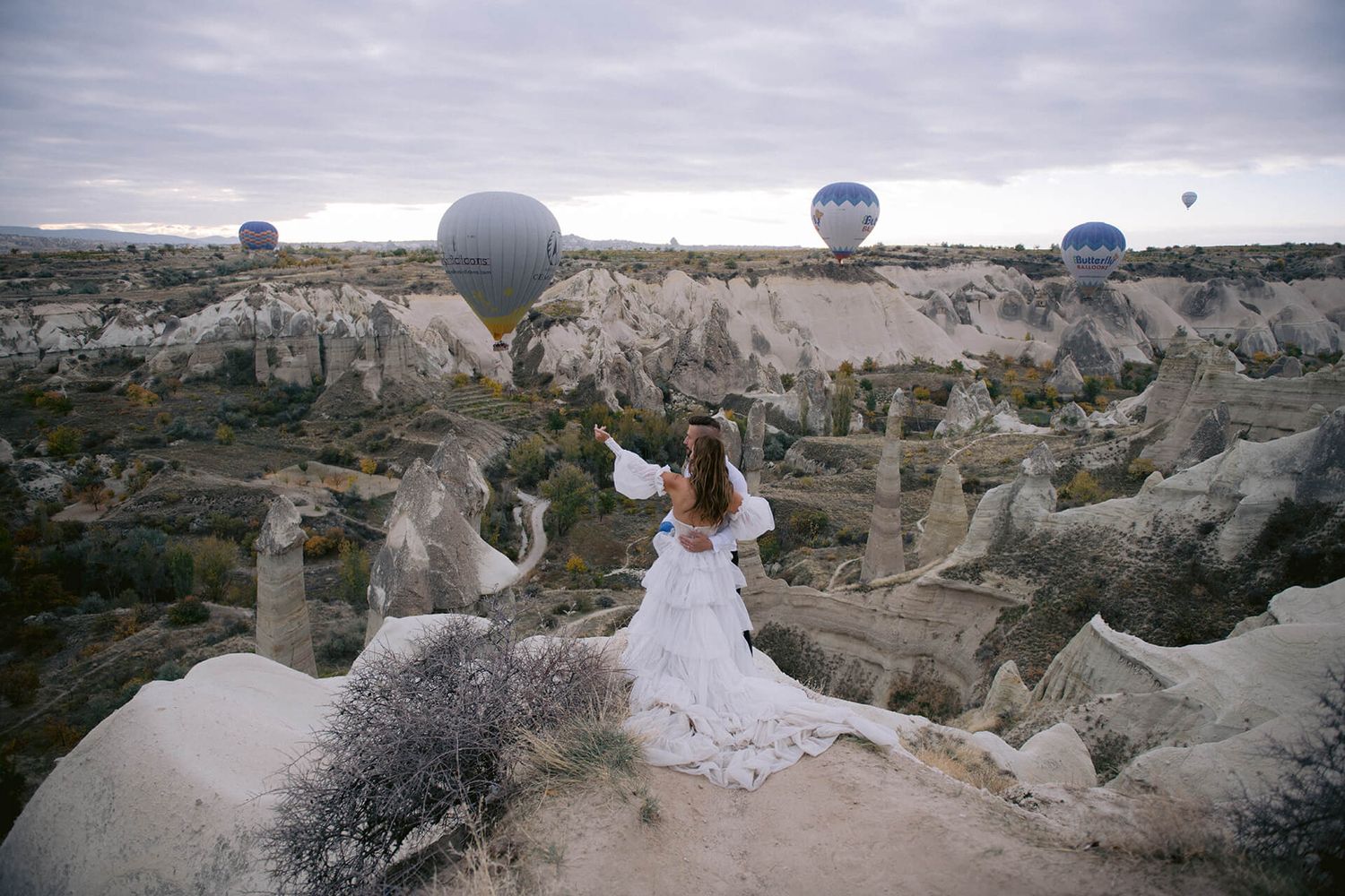 Karšto oro balionai virš Kapadokijos uolų debesuotą dieną – romantiška poros vestuvių fotosesijos „destination wedding“ atmosfera