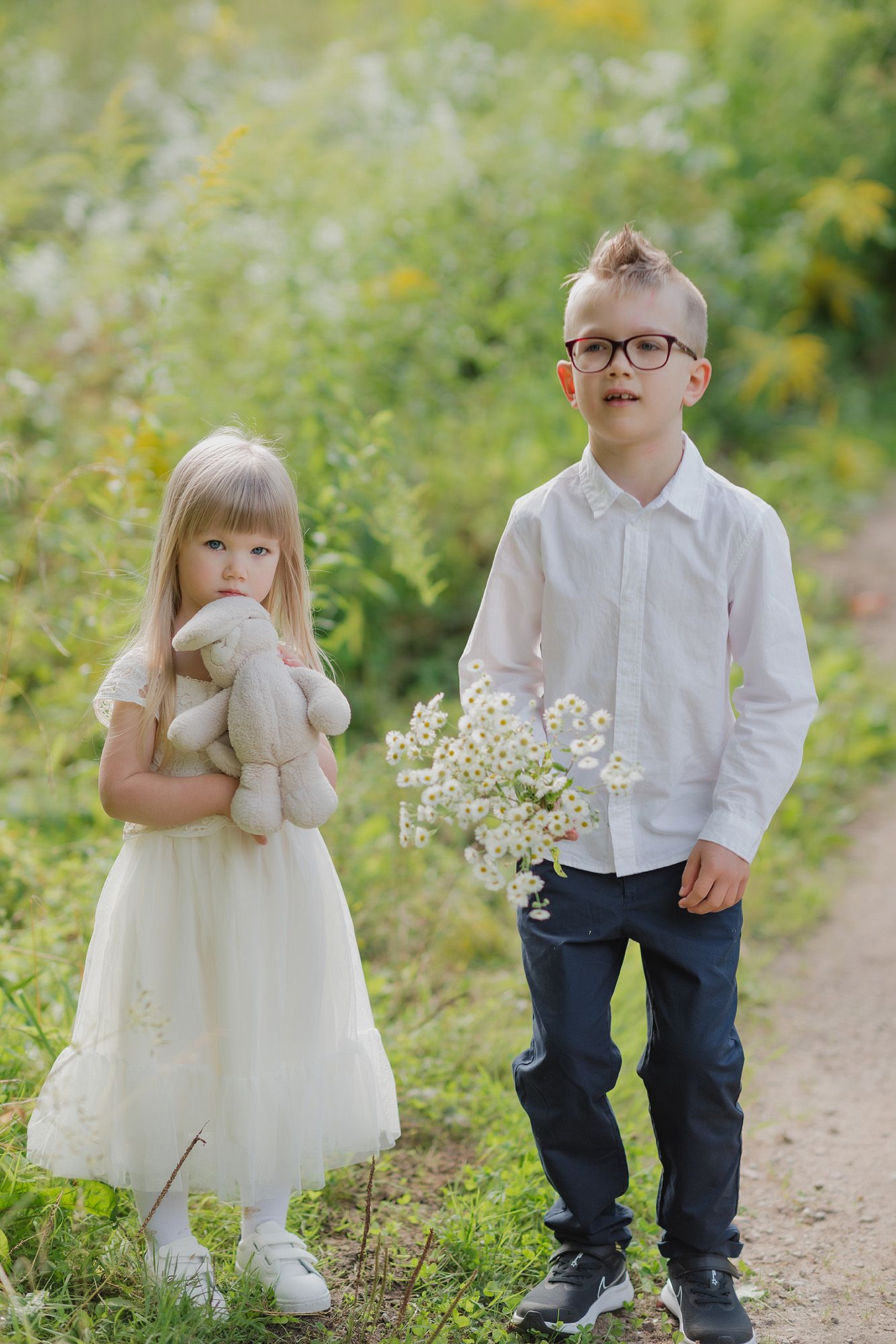 Two children stand on a garden path, one holding a stuffed toy and one holding wildflowers on a sunny summer day.