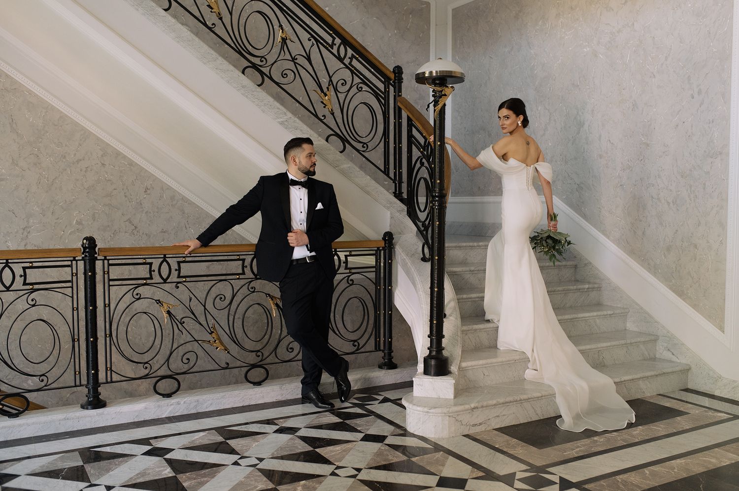A couple in formal wedding attire pose on an elegant marble staircase with ornate wrought iron railings.