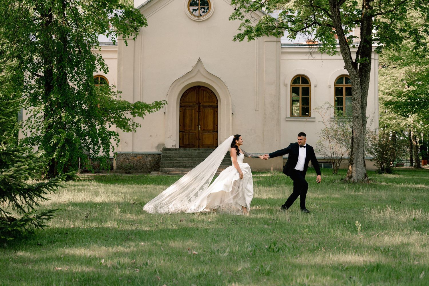 Bride and groom hold hands running across green lawn before a historic white church with arched wooden doors.