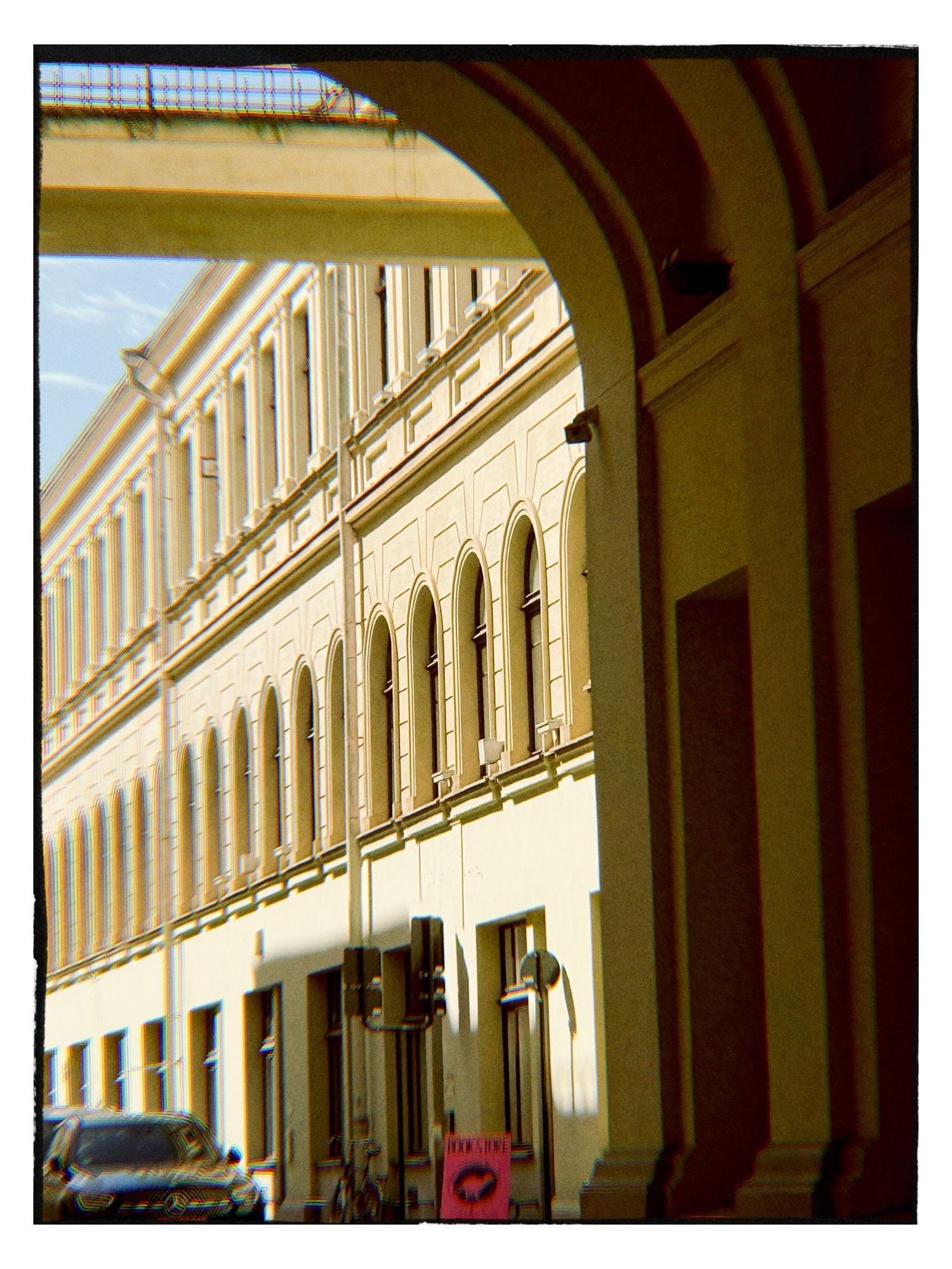 A sunlit European street viewed through an archway, featuring elegant yellow neoclassical buildings with arched windows.