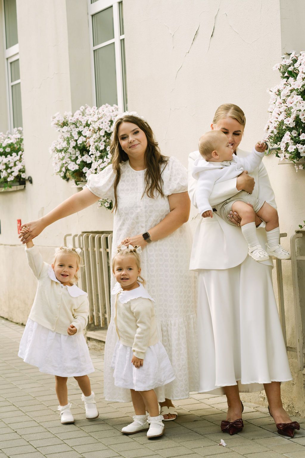 Two women in white dresses pose outdoors with three young children dressed in white near flower-adorned windows.