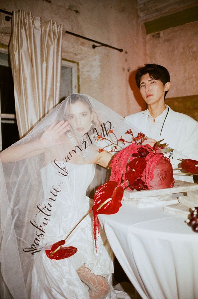 Bride in white veil and gown sits beside groom at table with red floral cake at elegant wedding.