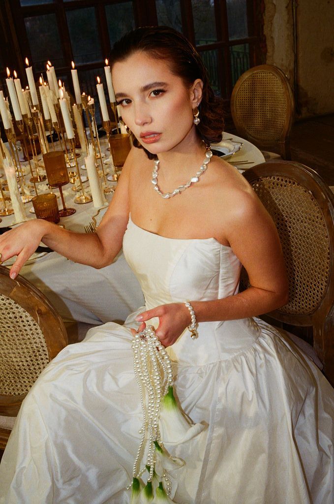Bride in strapless white gown seated at candlelit table, wearing pearl necklace and bracelet, holding pearl bouquet.