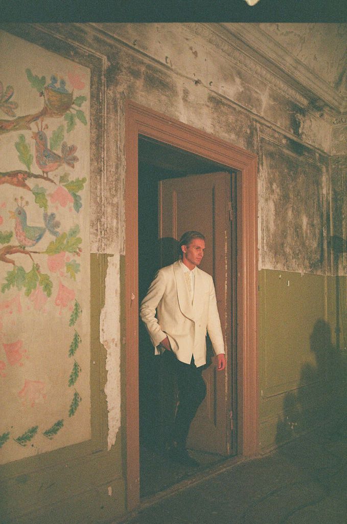 Man in white blazer standing in doorway of aged building with decorative bird and floral wall murals.