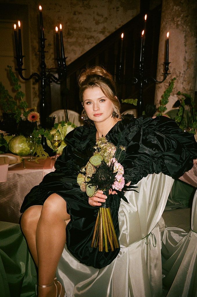 Young woman in black ruffled dress holding floral bouquet seated at elegant candlelit table with greenery decor.