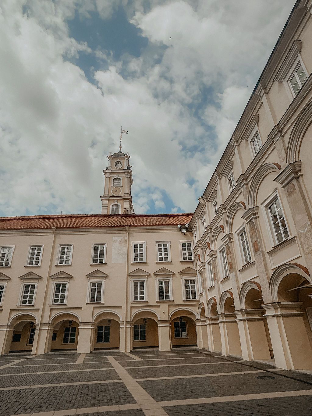 Historic university courtyard with arched colonnades, clock tower, and white baroque facade under a cloudy sky.