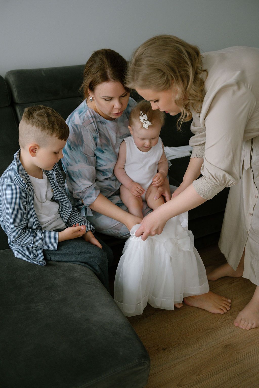 Two women and a young boy admire a baby girl in a white tulle dress on a gray sofa.
