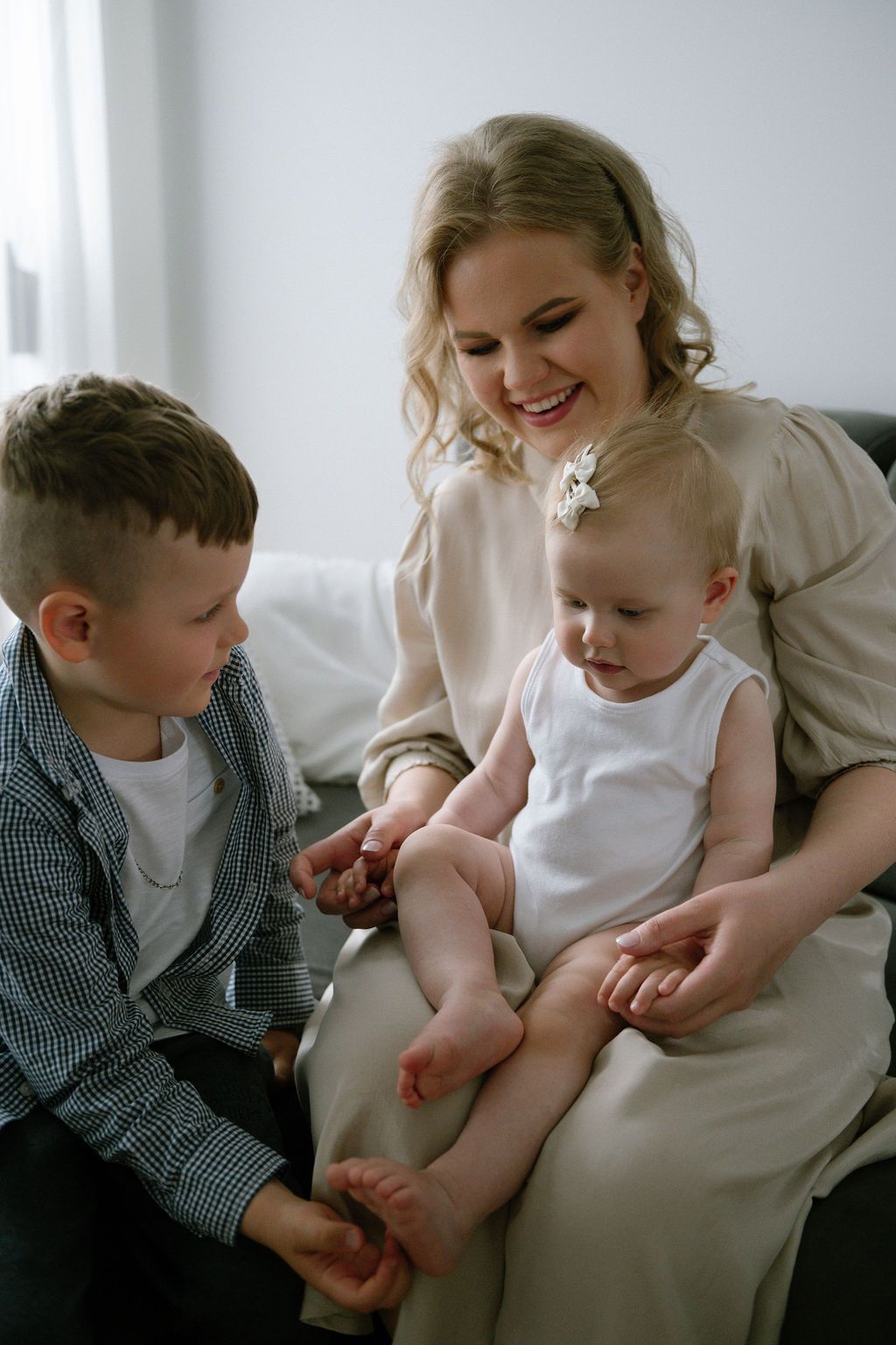 A smiling mother holds her baby while her young son looks on in a bright, cozy room.