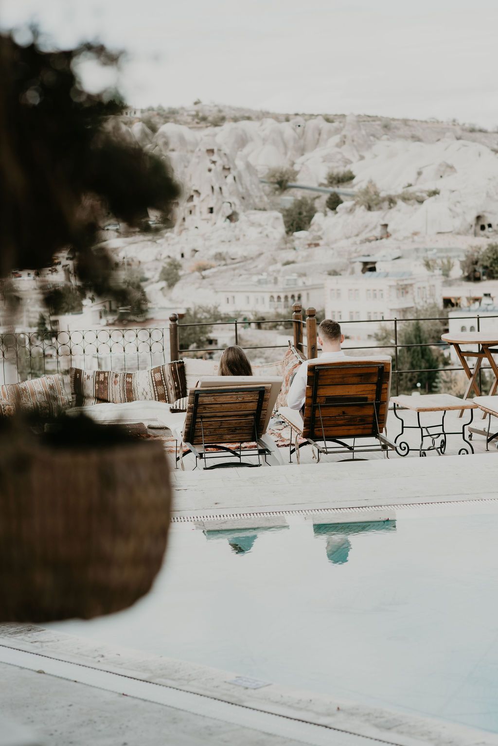 Couple relaxing on wooden lounge chairs by a pool overlooking the iconic cave rock formations of Cappadocia, Turkey.