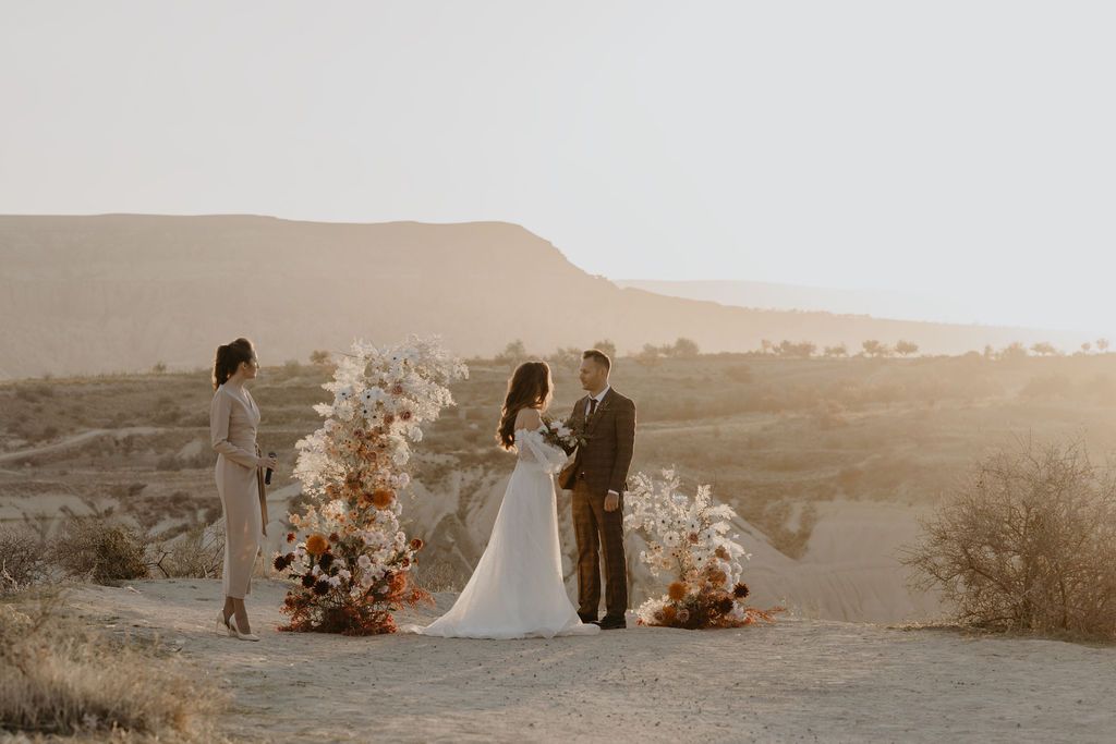 Boho desert wedding ceremony at sunset with bride, groom, and officiant surrounded by pampas grass and floral arrangements.