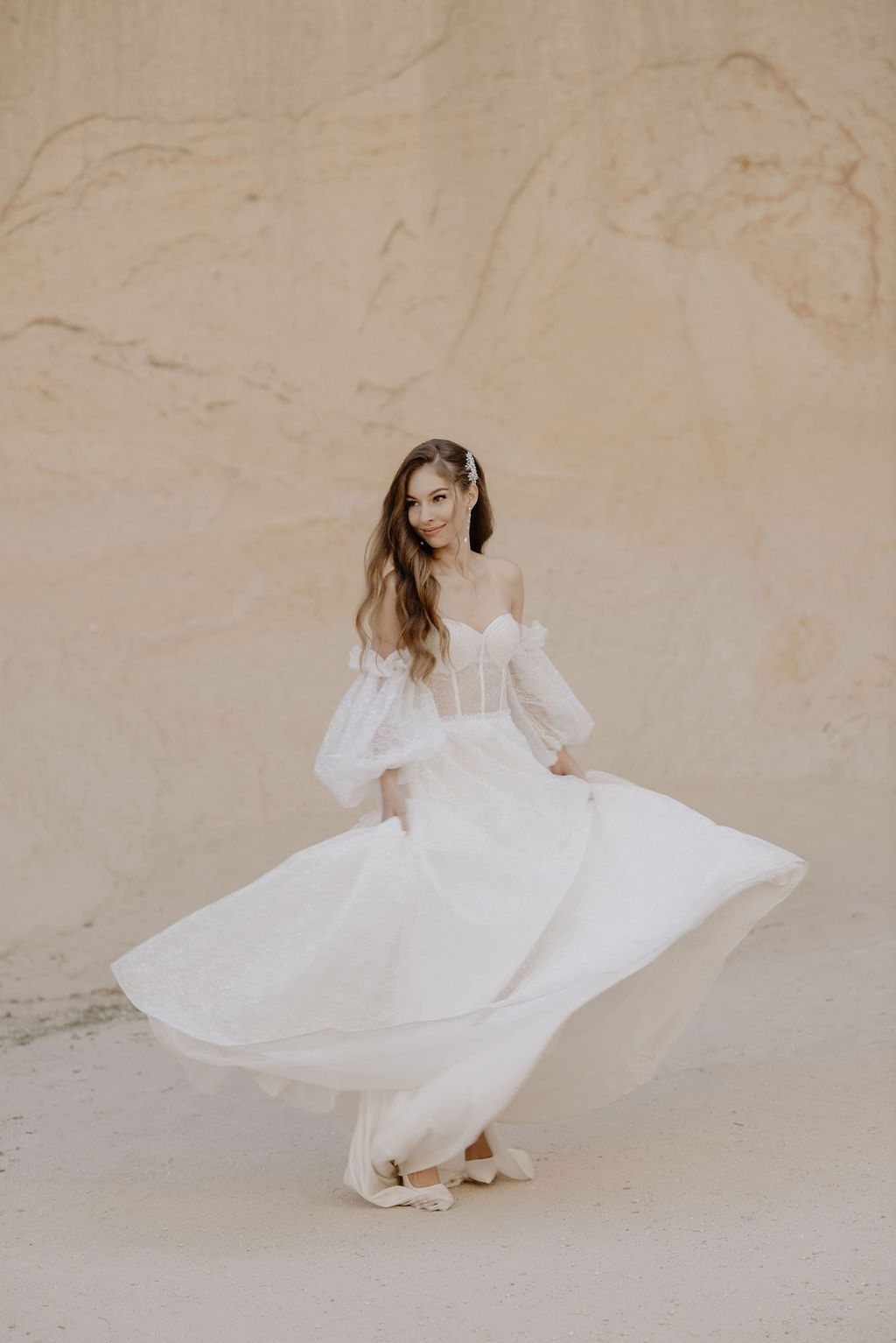 A bride twirls in a flowing white off-shoulder gown with puff sleeves against a warm sandy stone wall.
