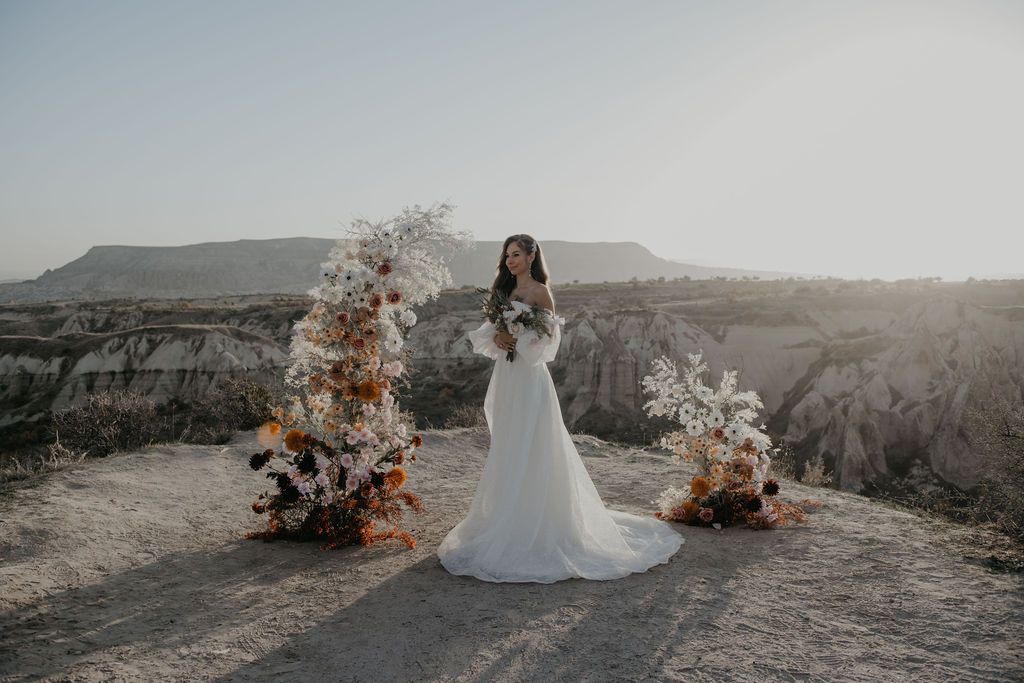 Bride in white gown standing between floral arrangements with canyon views at sunset.