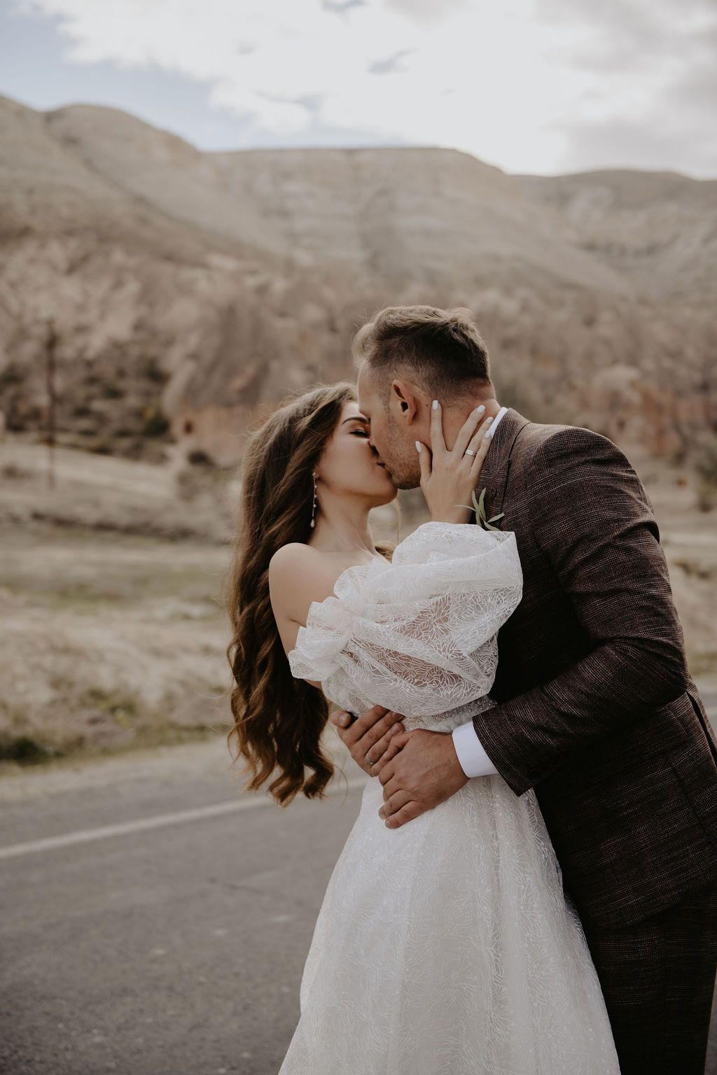 A bride and groom share a romantic kiss outdoors with scenic desert mountains in the background on their wedding day.