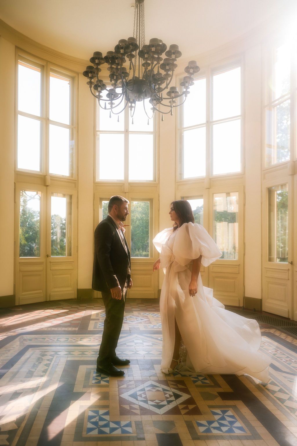 Bride in puffy sleeve gown and groom in tuxedo share a moment in sunlit ballroom with ornate chandelier.