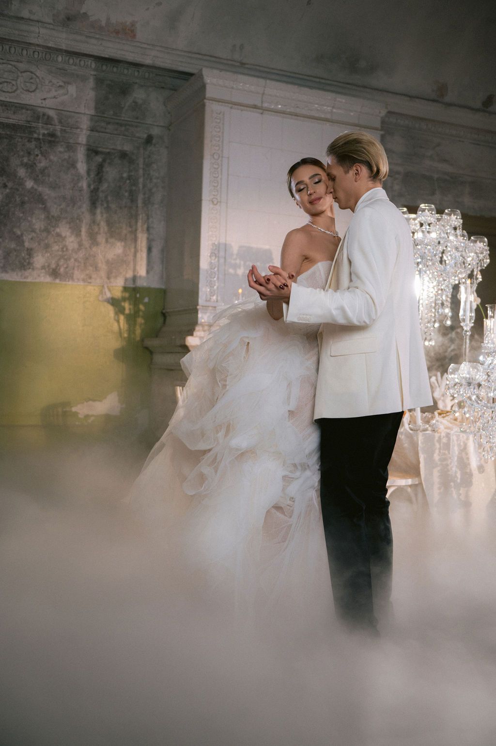 Bride and groom share first dance surrounded by fog in elegant venue, bride in white gown, groom in white tuxedo jacket.