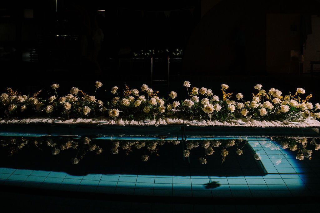 Elegant floral arrangement of white roses reflected in a pool at a dark, sophisticated indoor event.