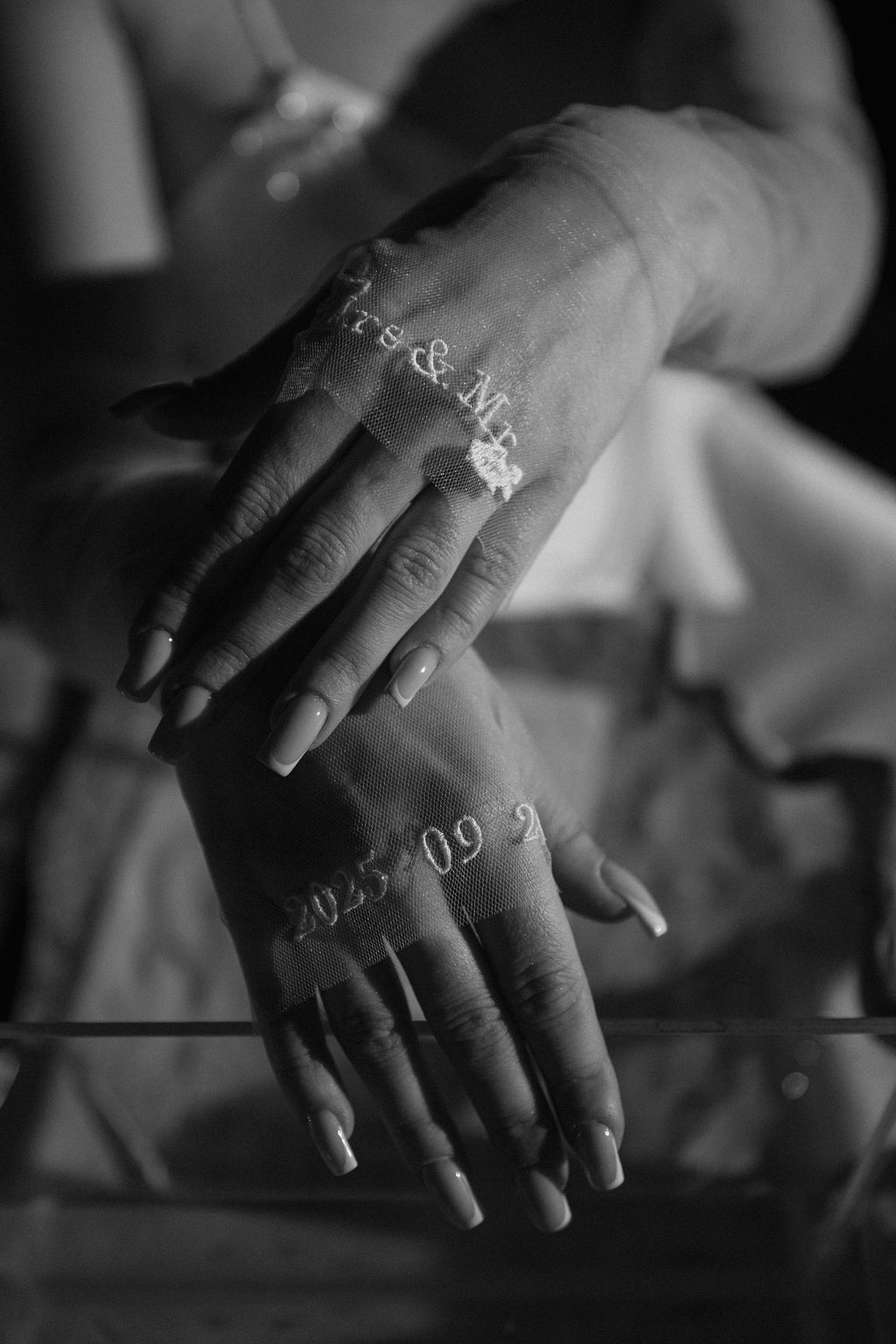 Black and white photo of bride's hands with French manicure resting on glass surface with Mrs & Mr wedding date reflection.