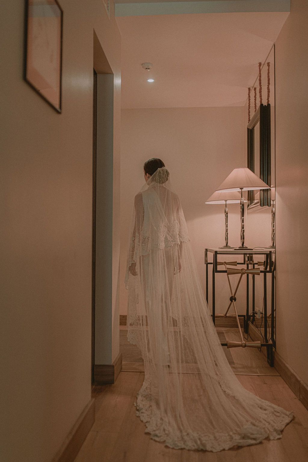 Bride in elegant white lace wedding gown with long cathedral veil standing in softly lit hotel corridor.
