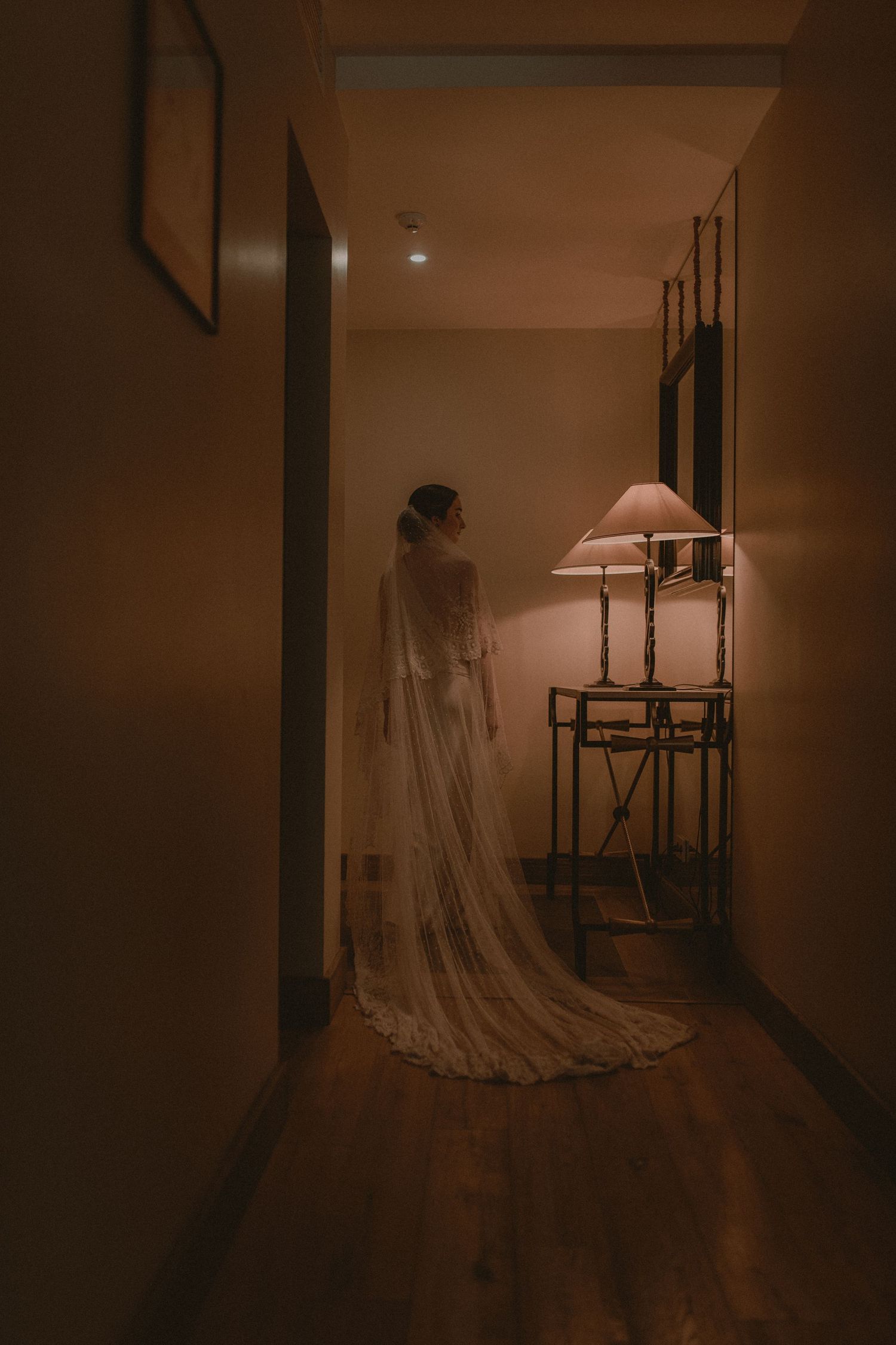 Bride in a flowing white wedding gown with long veil stands in a dimly lit hallway beside a warm glowing lamp.