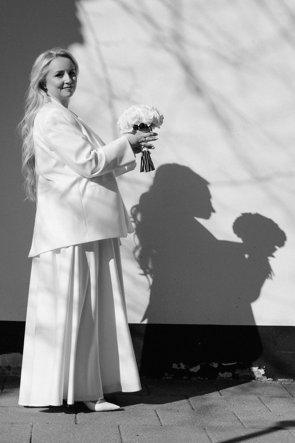 Blonde bride in white suit holds bouquet casting dramatic shadow on wall, black and white wedding photo.