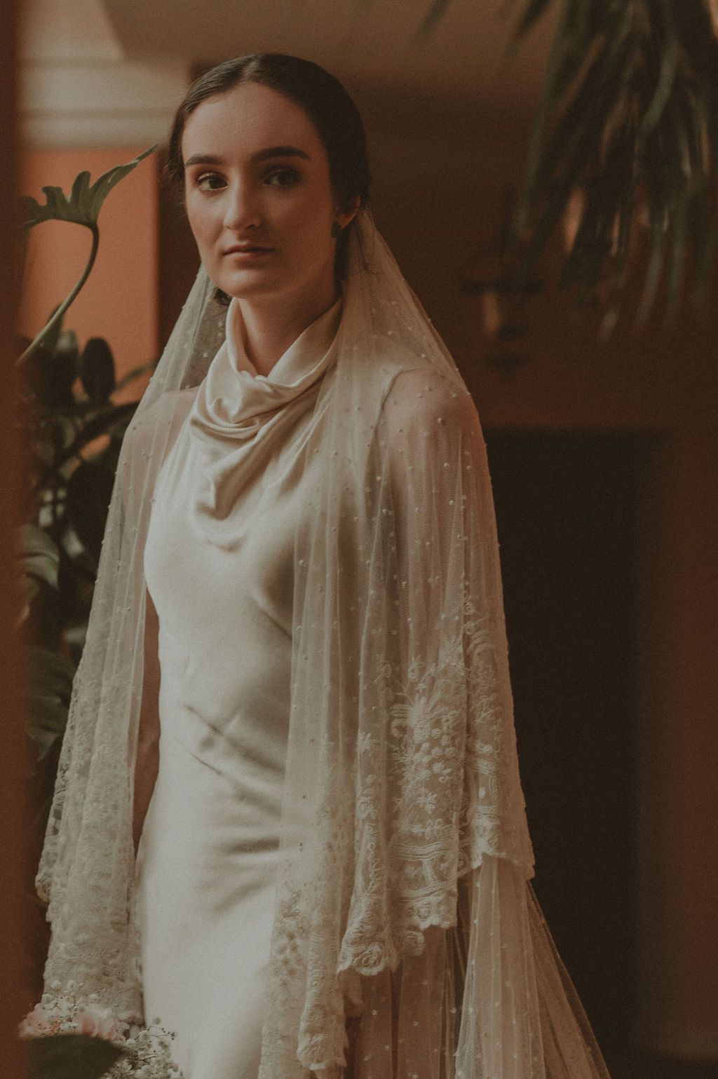 Elegant bride wearing a beaded lace veil and satin wedding dress in a warm-toned interior.