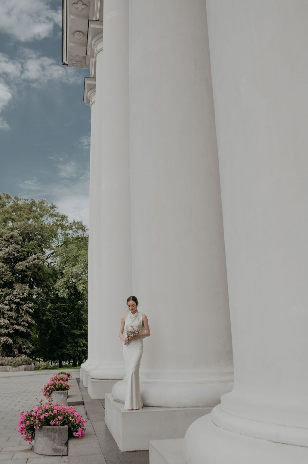 Bride in white gown standing between large white columns of elegant building with pink flowers and trees in background.