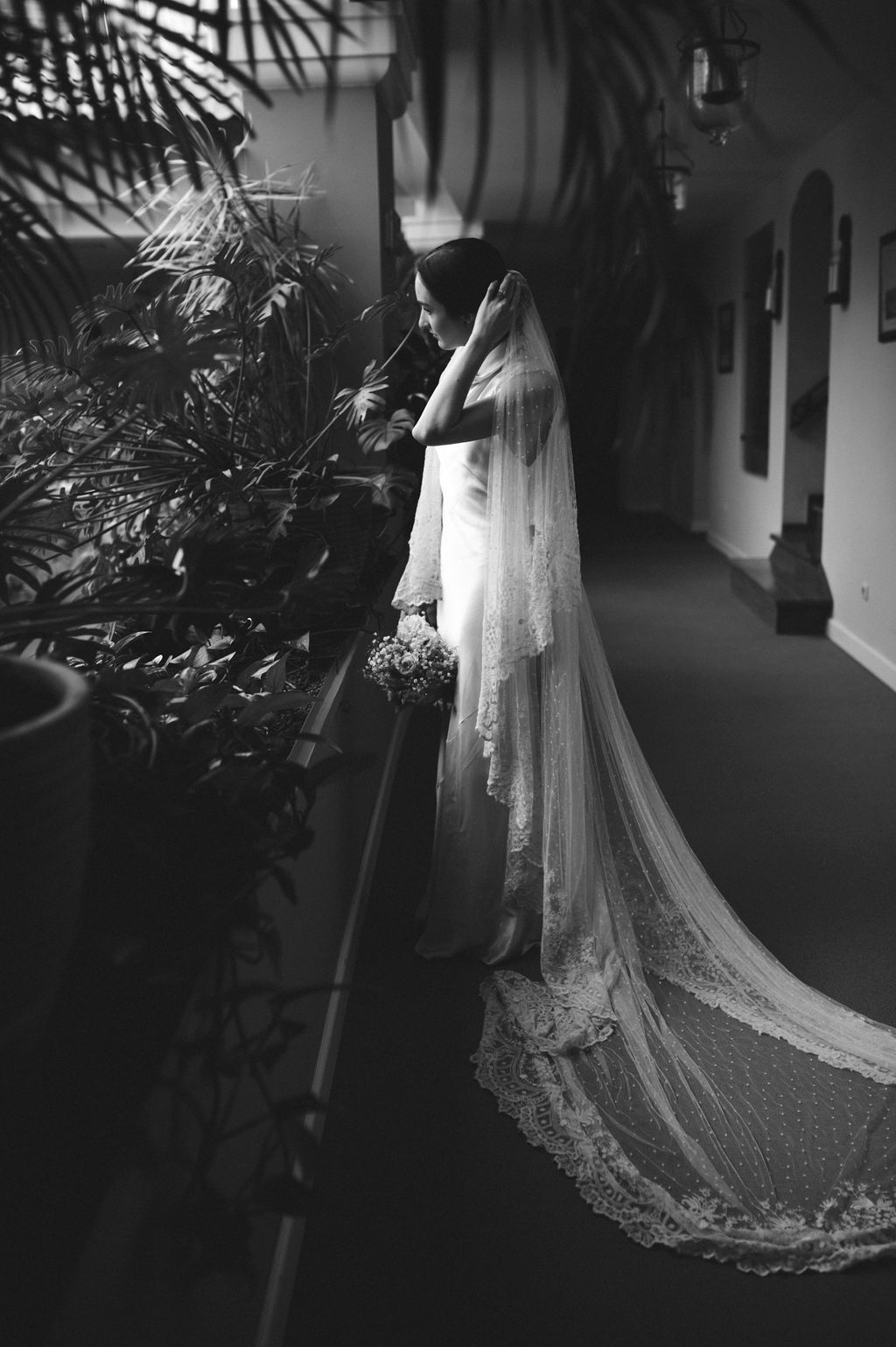 Black and white photo of bride in a flowing gown and long lace veil standing among tropical plants.