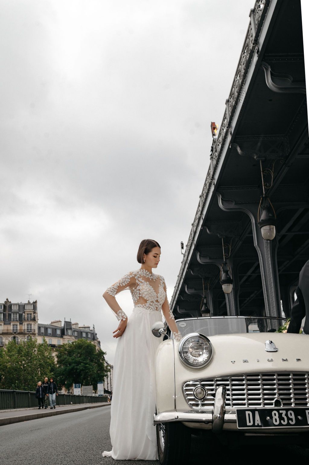 Bride in white lace gown poses beside vintage cream car on Parisian street under cloudy sky.