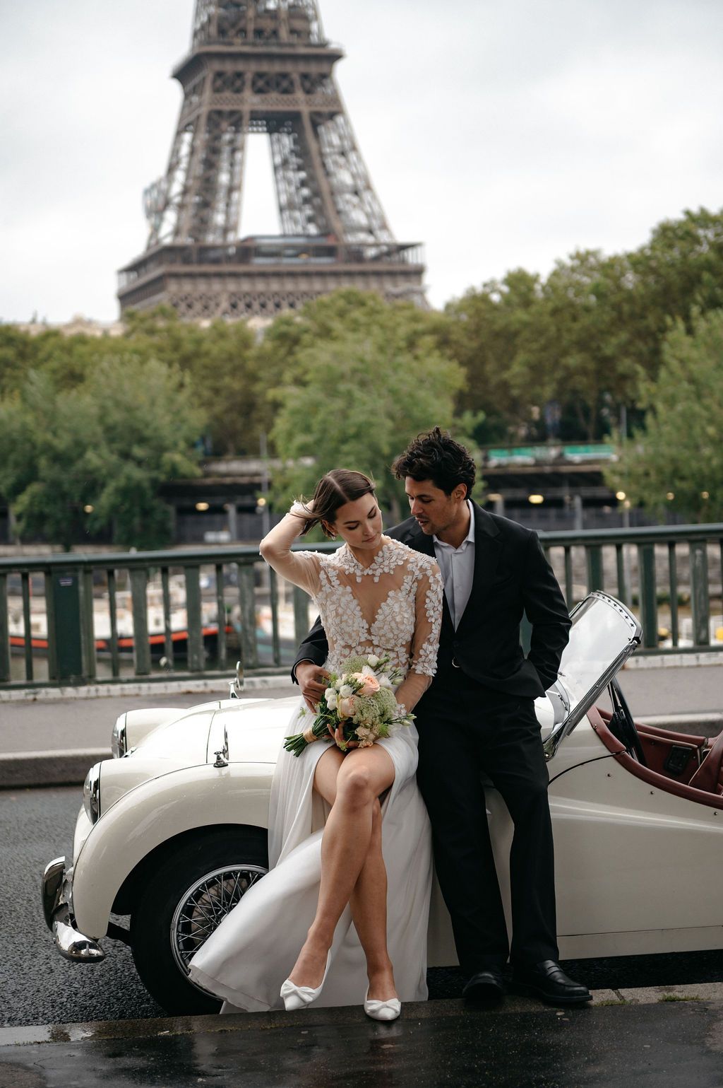 Bride and groom pose with vintage white convertible car near Eiffel Tower in Paris on wedding day.