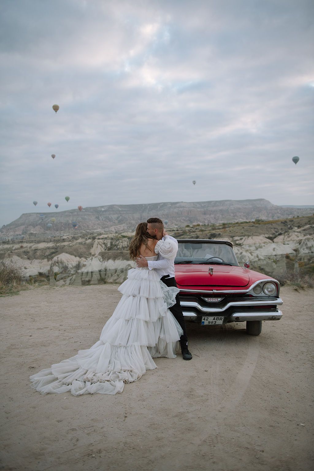 Bride and groom kissing beside a vintage red convertible in Cappadocia with hot air balloons floating in the sky.