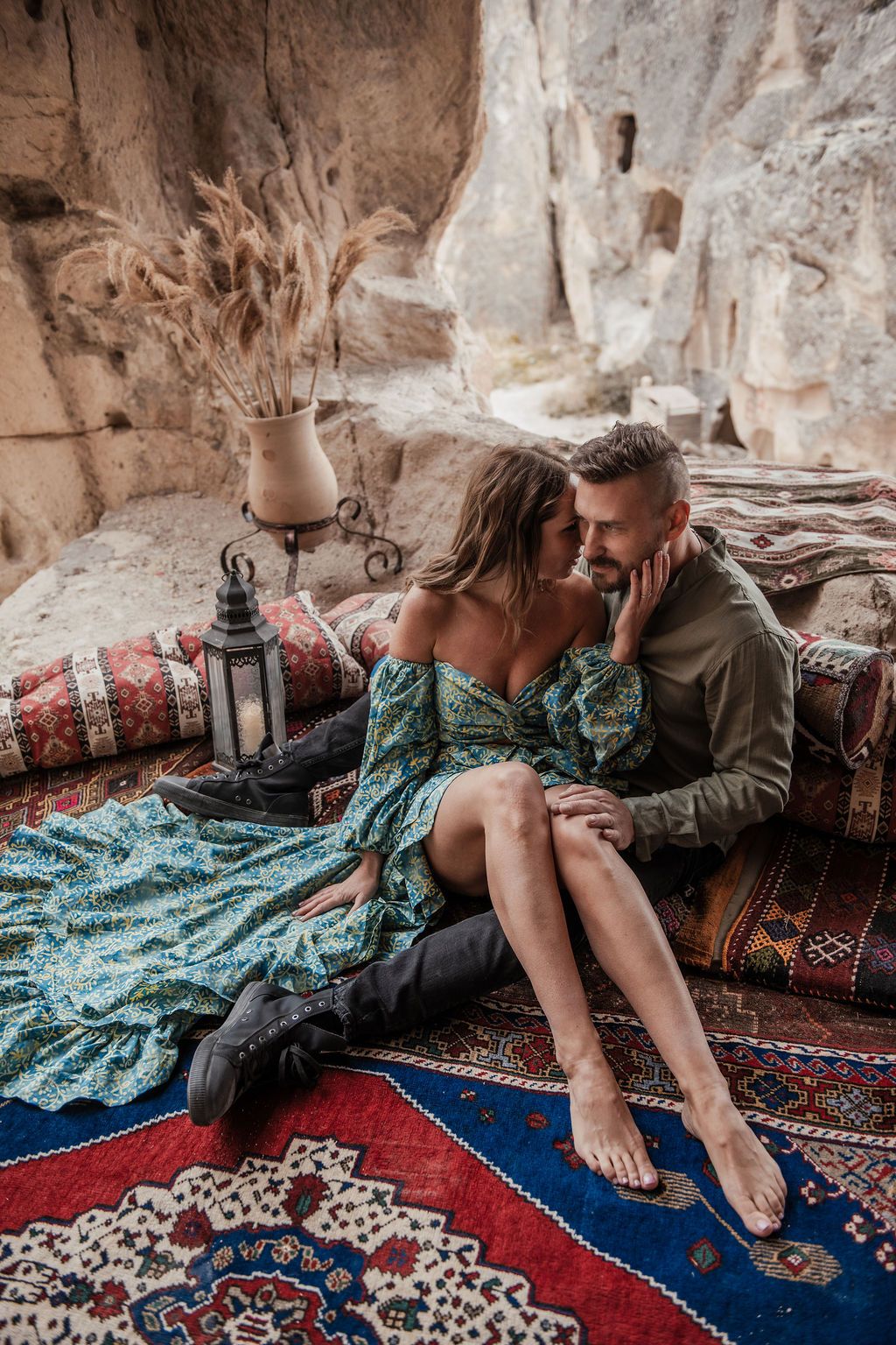 Romantic couple sitting on colorful rugs in a cave dwelling, woman in green floral dress with boots, man in olive shirt.