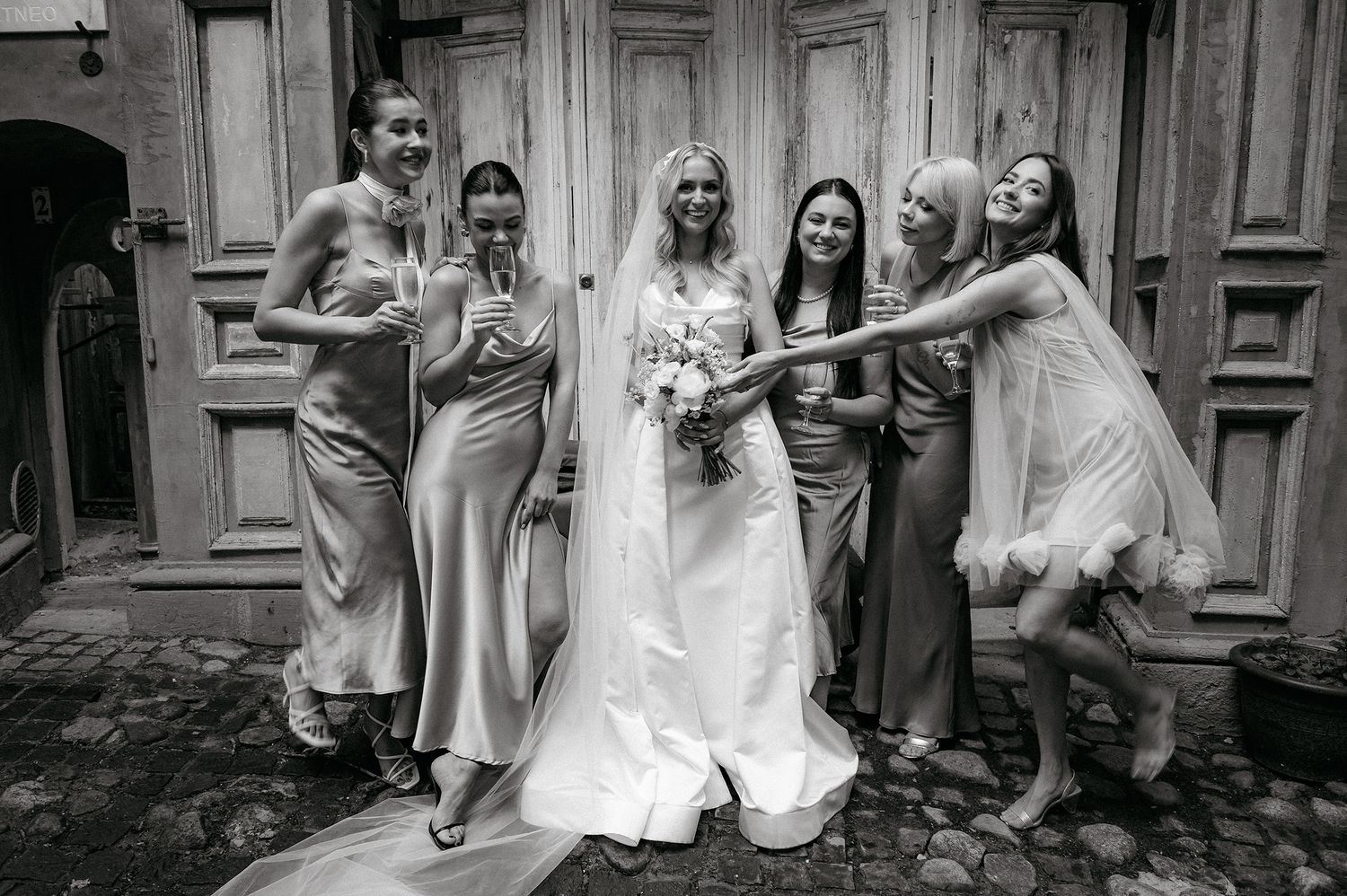 A wedding party poses together in black and white while making silly faces and gestures on a building stoop.