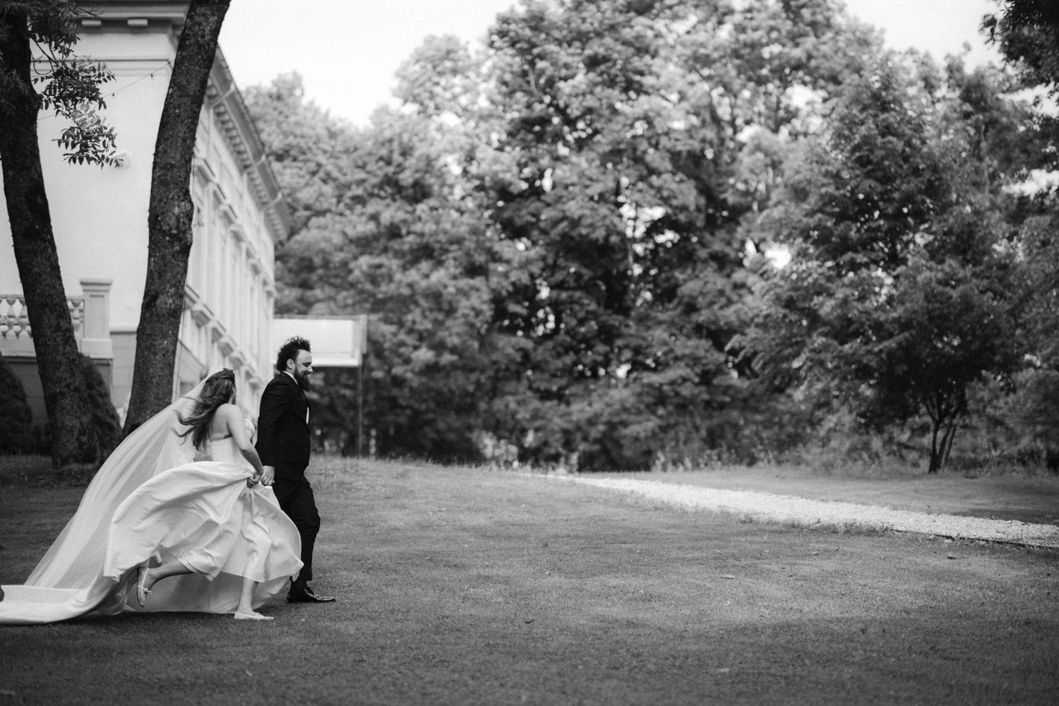 Black and white photo of a bride and groom walking outdoors near a building surrounded by lush trees.