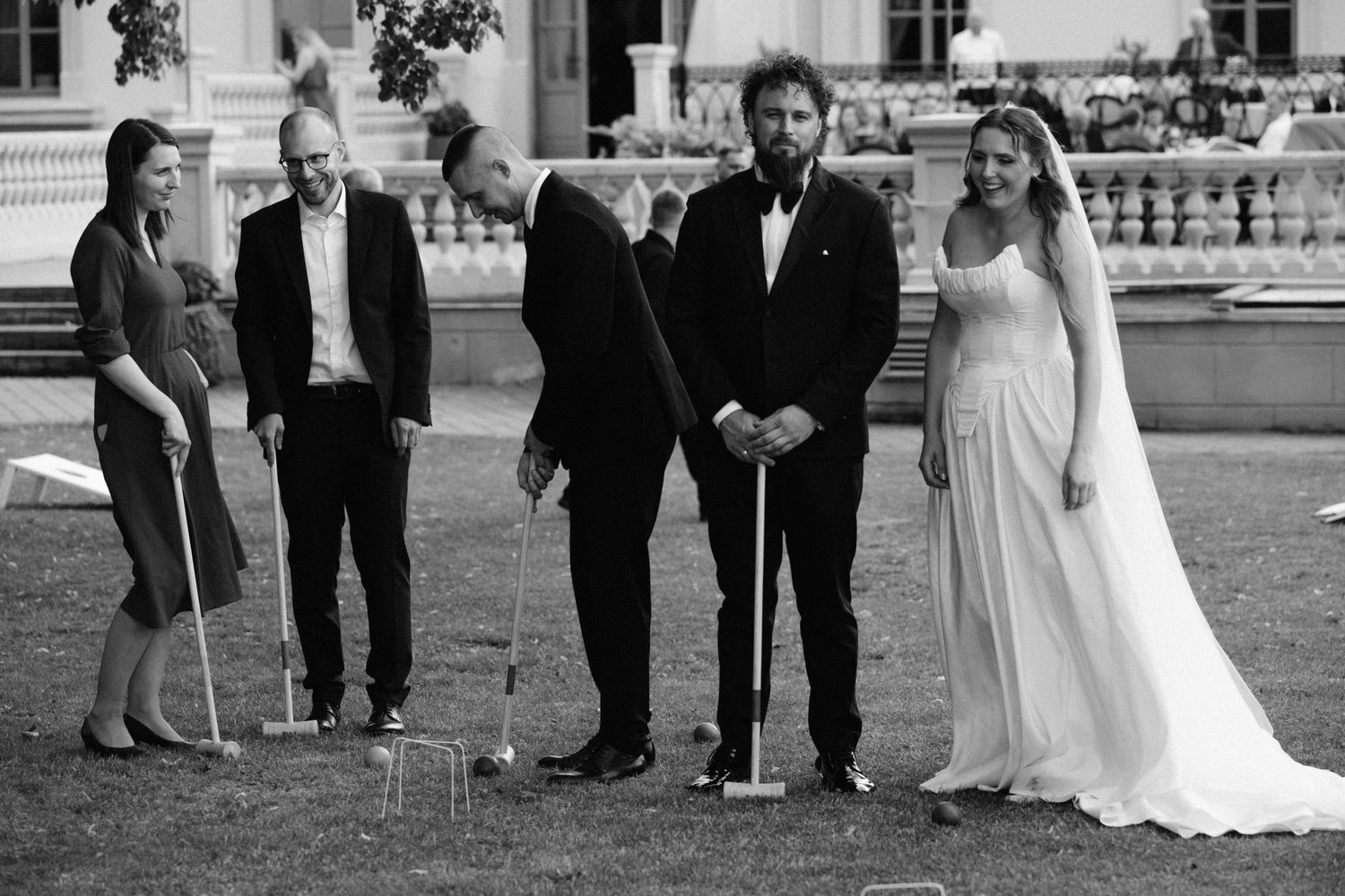Black and white photo of a bride and groom playing croquet with guests at an elegant outdoor wedding reception.