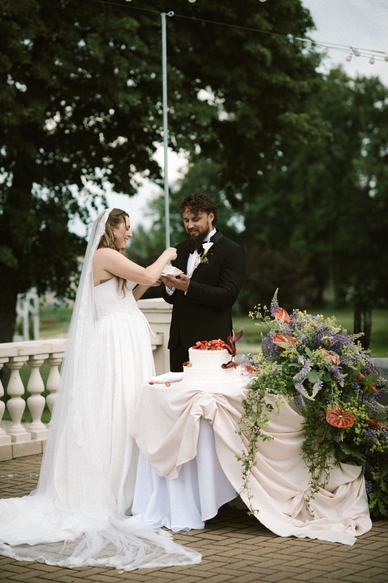 Bride and groom cutting their wedding cake outdoors surrounded by lush floral arrangements and greenery.