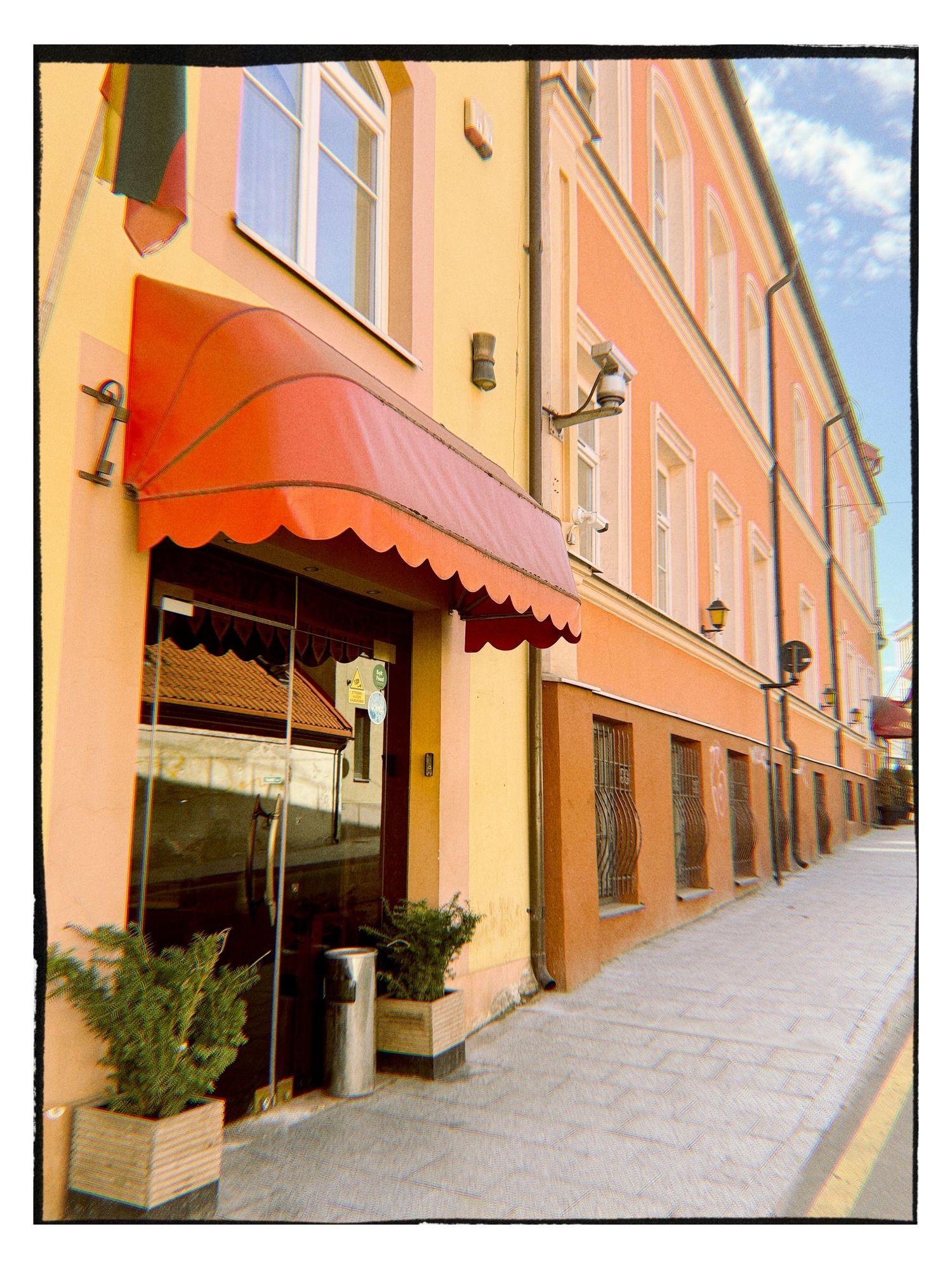 Orange building exterior with red scalloped awning over entrance, potted plants, and cobblestone sidewalk in European town.