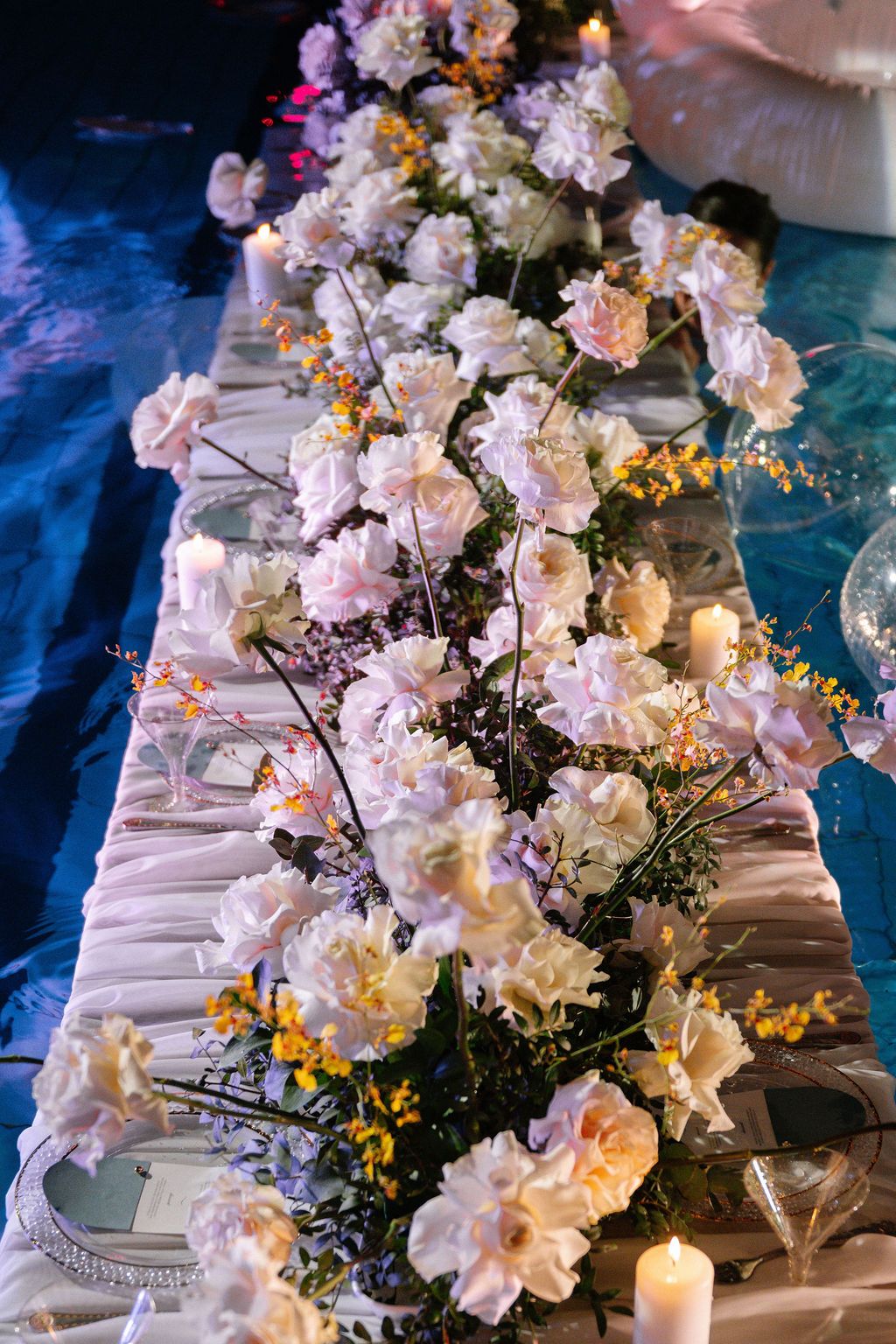 Long wedding reception table decorated with blush pink roses, orange accents, candles and ivory draping under soft blue lighting.